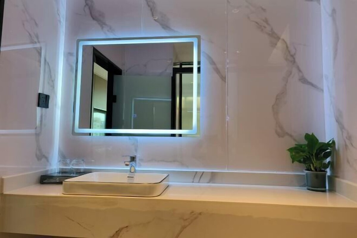 Une salle de bain avec un meuble-lavabo en marbre, un miroir rectangulaire doté d’un éclairage à LED et une plante en pot posée sur le plan de travail.