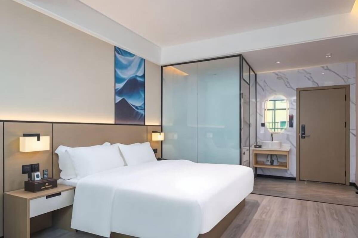 A hotel room with a large bed, bedside lamps, a nightstand, a glass partition, a bathroom with a sink, and a door.