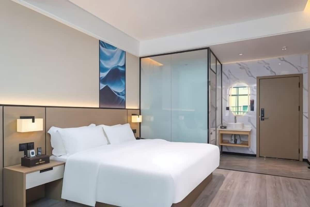 A hotel room with a large bed, bedside lamps, a nightstand, a glass partition, a bathroom with a sink, and a door.