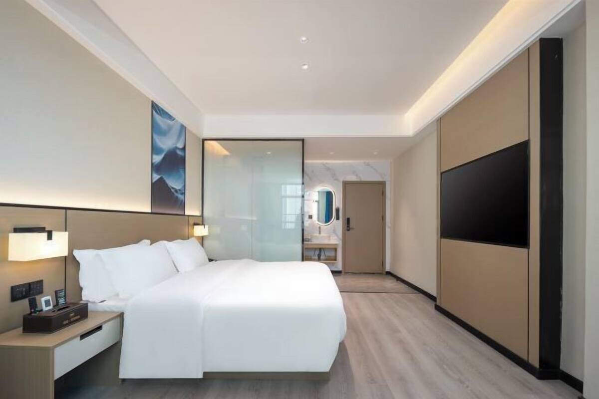 A modern hotel room with a large bed, a bedside table with a lamp, a flat-screen TV, and a large window.