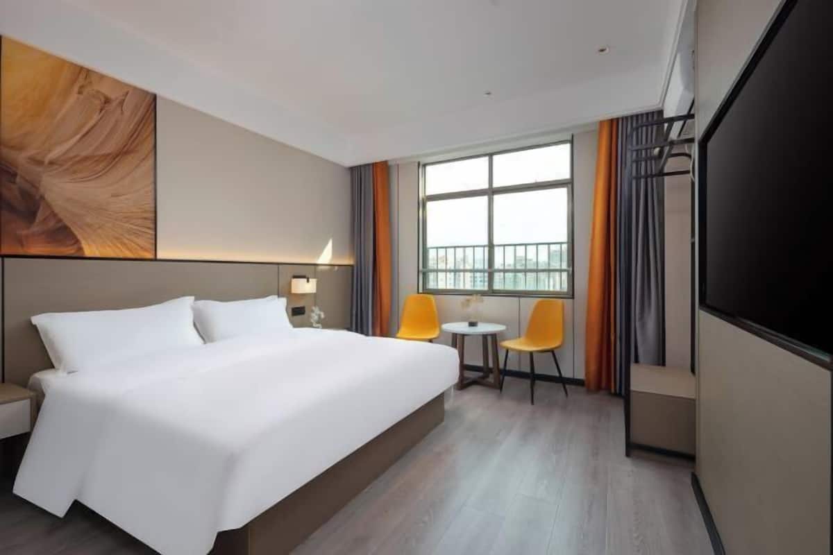 A hotel room with a large bed, two chairs, a small table, a TV, and a large window.