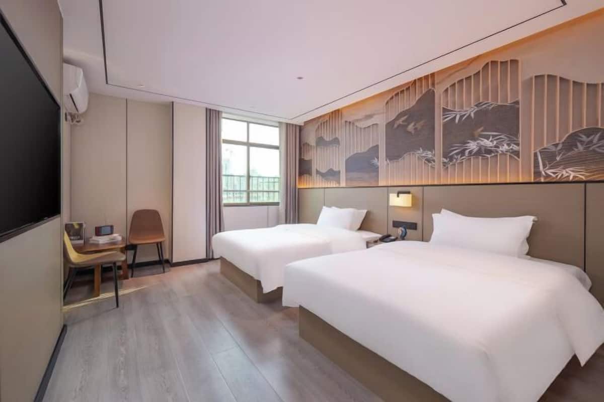 A hotel room with two beds, a desk with a chair, a TV, and a large window.