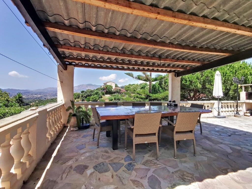 Une terrasse extérieure avec un sol en pierre, une table en bois, des chaises en osier et une pergola au-dessus.