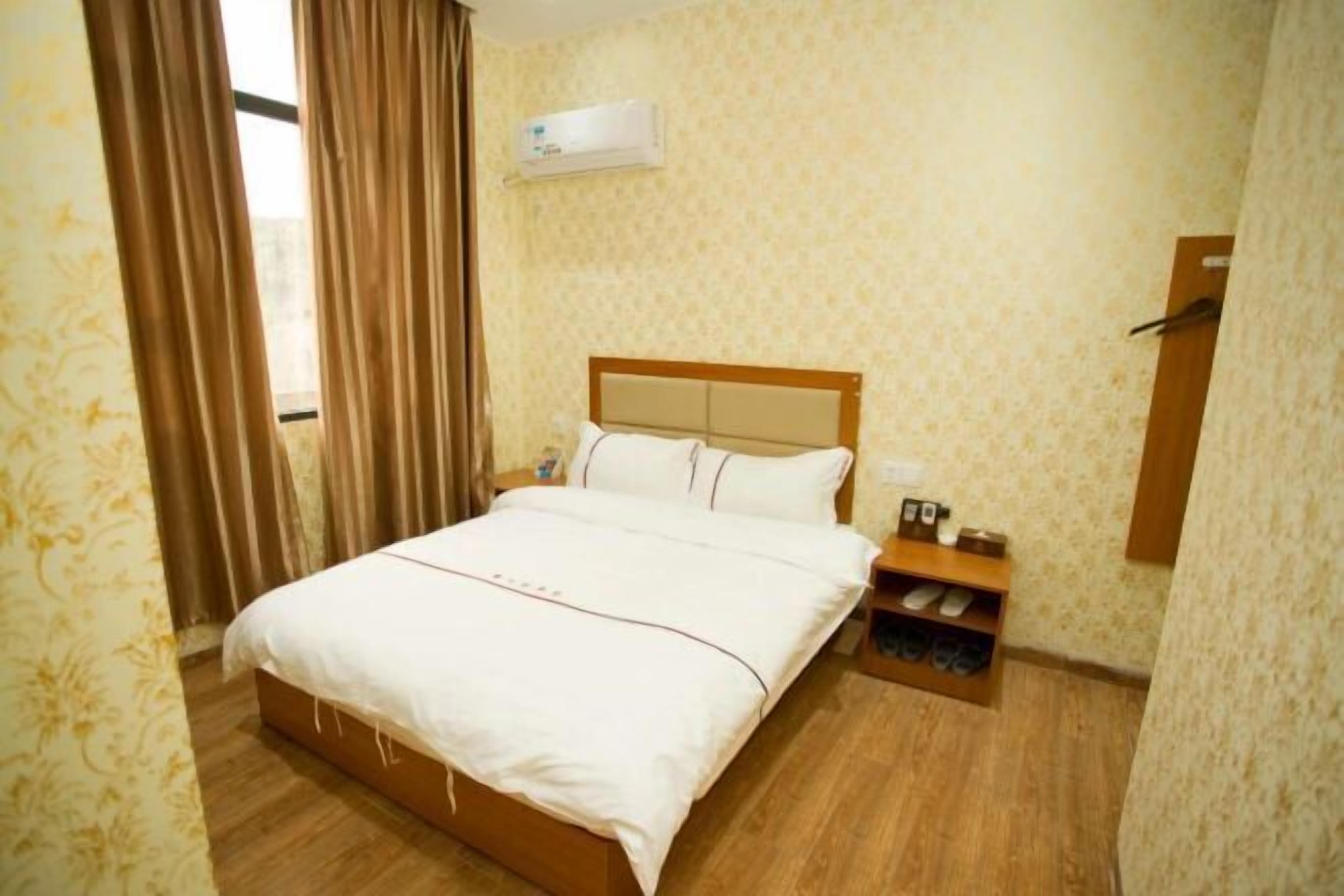 A hotel room with a bed, a nightstand, a shoe rack, and an air conditioning unit.