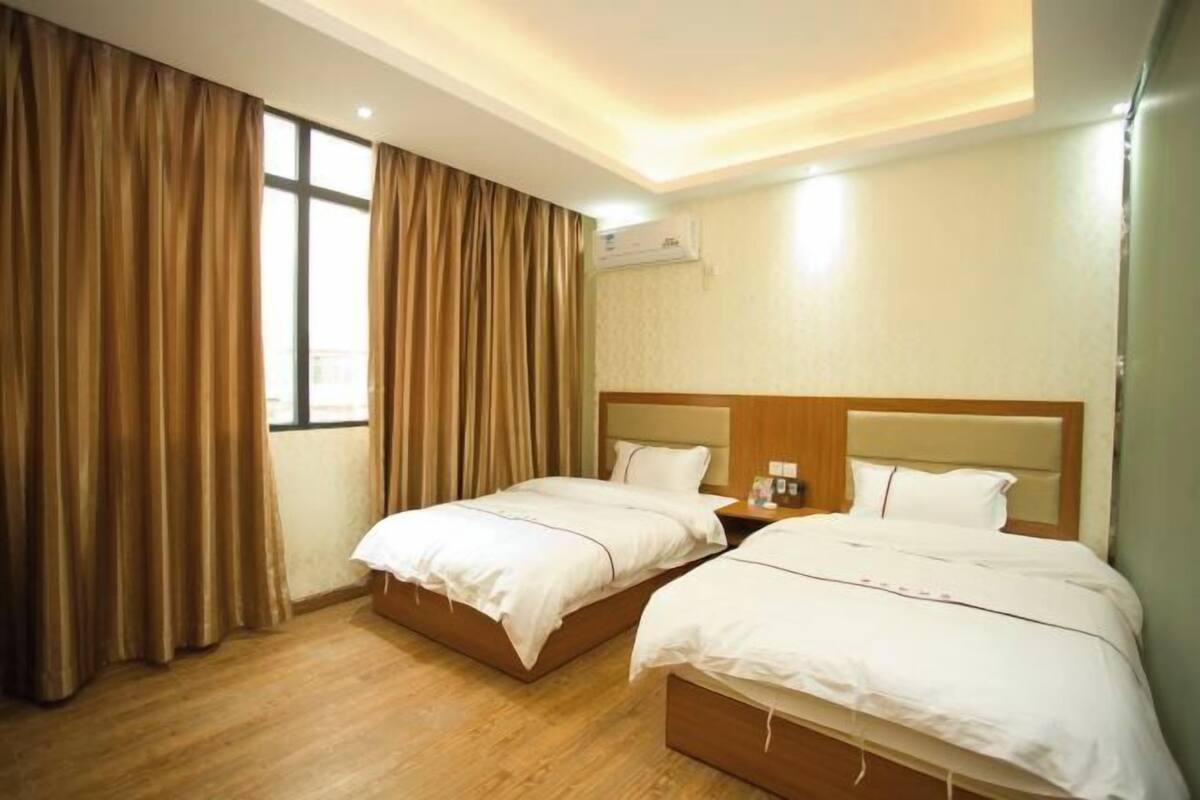 A hotel room with two beds, a window with curtains, an air conditioning unit, and a wooden floor.