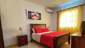 A neatly arranged bedroom with a bed, a nightstand, a lamp, a wall-mounted air conditioner, and a window with curtains.