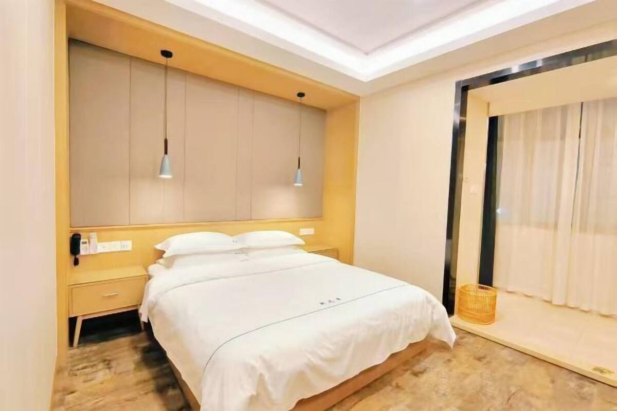 A hotel room with a bed, bedside table, lamp, wardrobe, and a window with curtains.