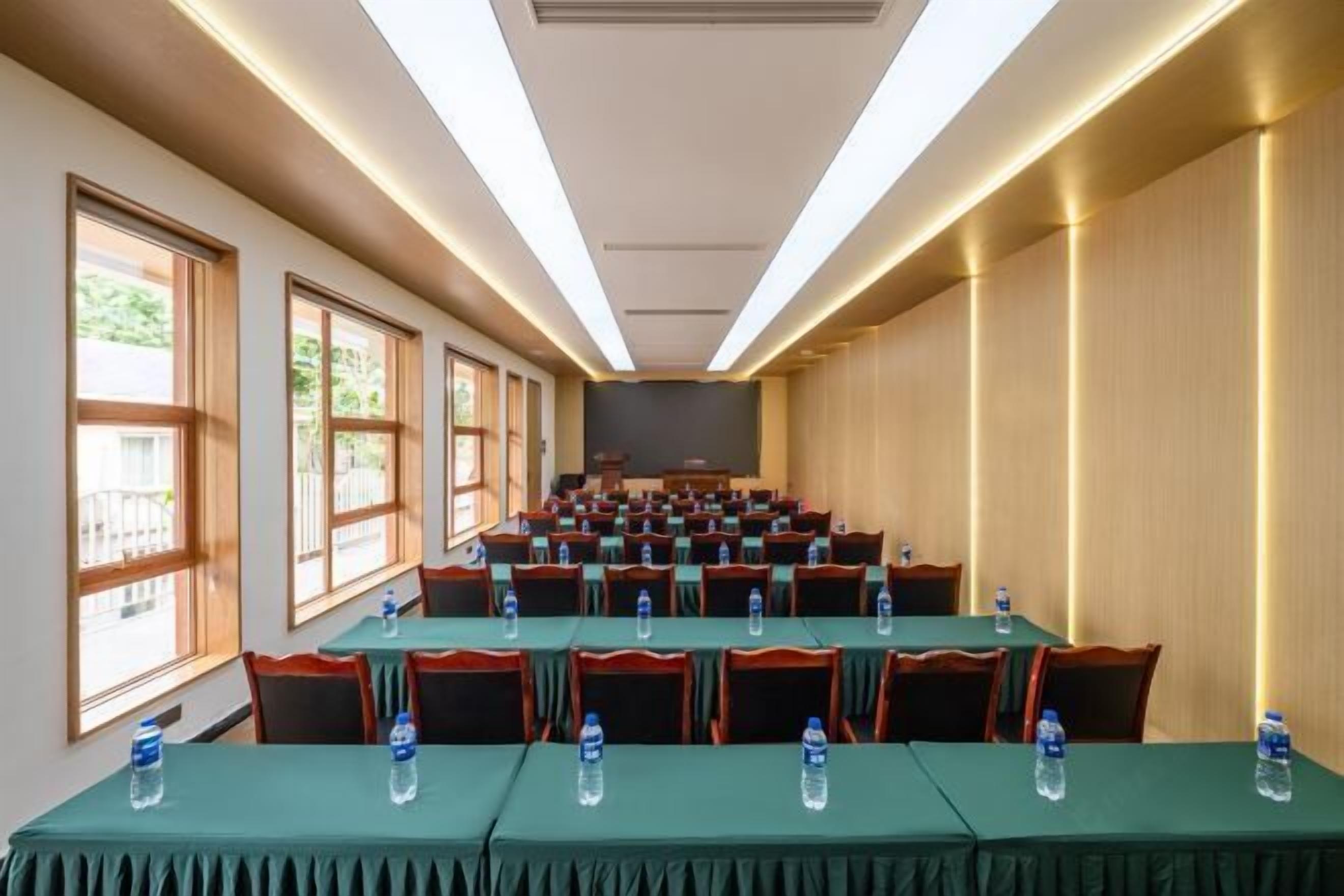 Un salón de conferencias con filas de mesas y sillas, equipado con botellas de agua y grandes ventanales en un lateral.