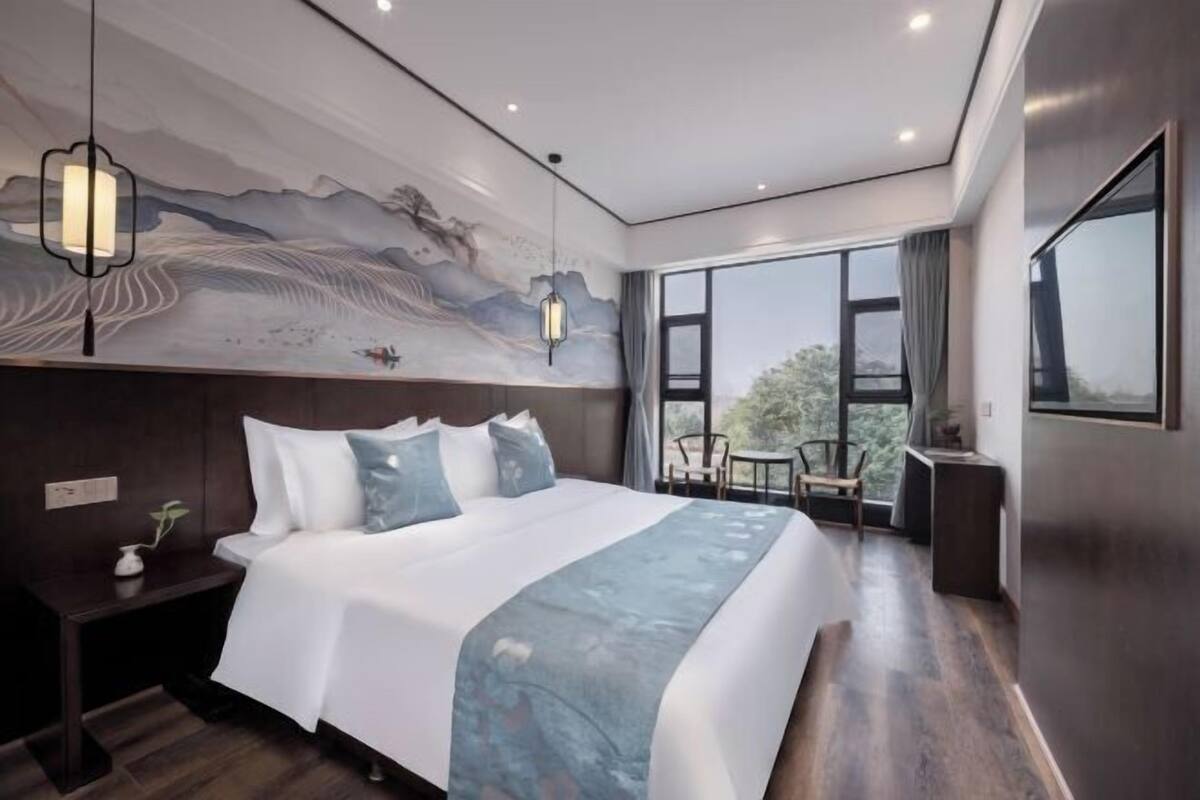 A modern hotel room with a large bed, a mural on the wall, a view through the window, and a small table with a chair.