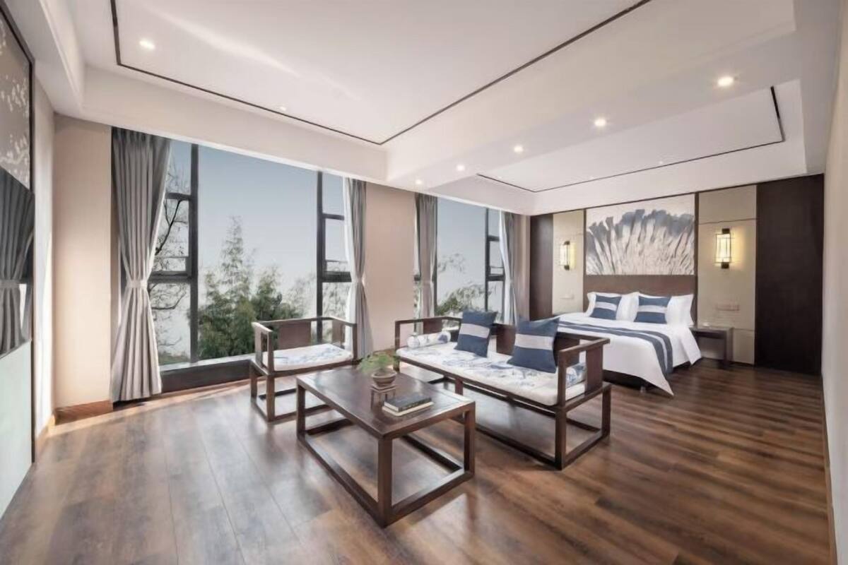 A modern hotel room with a large bed, a seating area with a table, and large windows offering a view of trees.