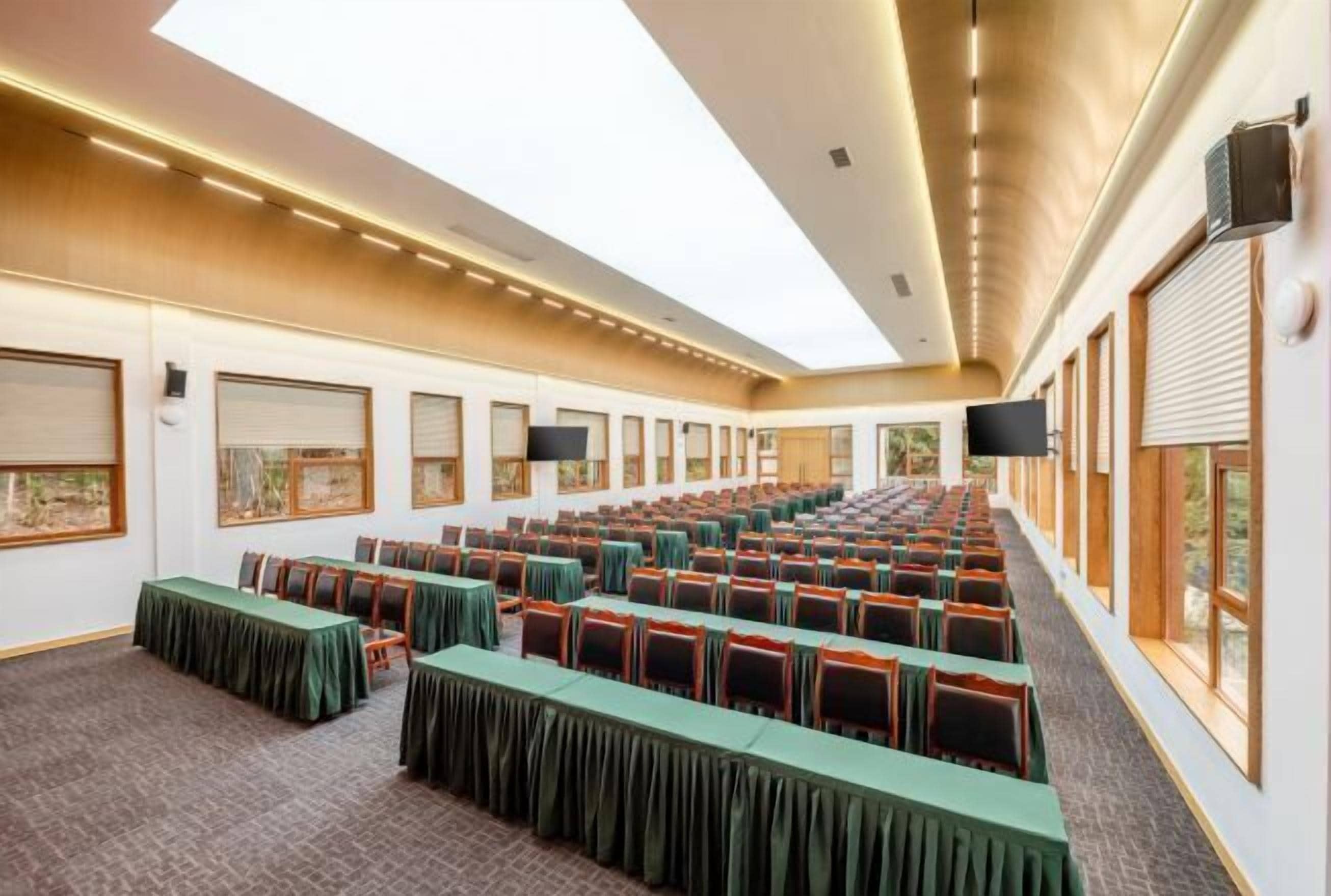A conference room with rows of tables and chairs, large windows, and a high ceiling with recessed lighting.