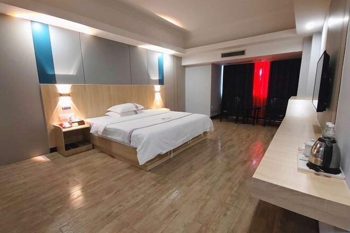 A modern hotel room with a wooden floor, a bed with white linens, a bedside table with a lamp, a small shelf, and a minibar.