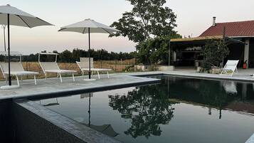 A swimming pool with lounge chairs and umbrellas, surrounded by trees and a building with a red roof.