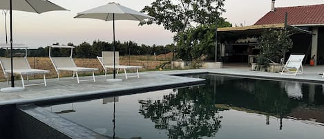 A swimming pool with lounge chairs and umbrellas, surrounded by trees and a building with a red roof.