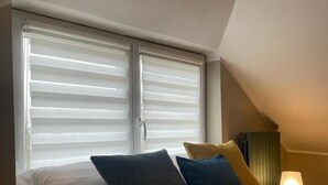 Une chambre à coucher avec un lit, des oreillers, une fenêtre avec des stores et une table de chevet avec une lampe.