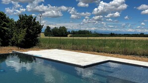 Une piscine bordée de béton, entourée d’un champ verdoyant et d’arbres, sous un ciel partiellement nuageux.