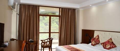 A hotel room with a large bed, a desk, and a chair. There is a window with curtains, and the room has a ceiling light.