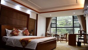 Quarto de hotel com cama, escrivaninha, cadeiras e uma janela grande com vista para o verde.