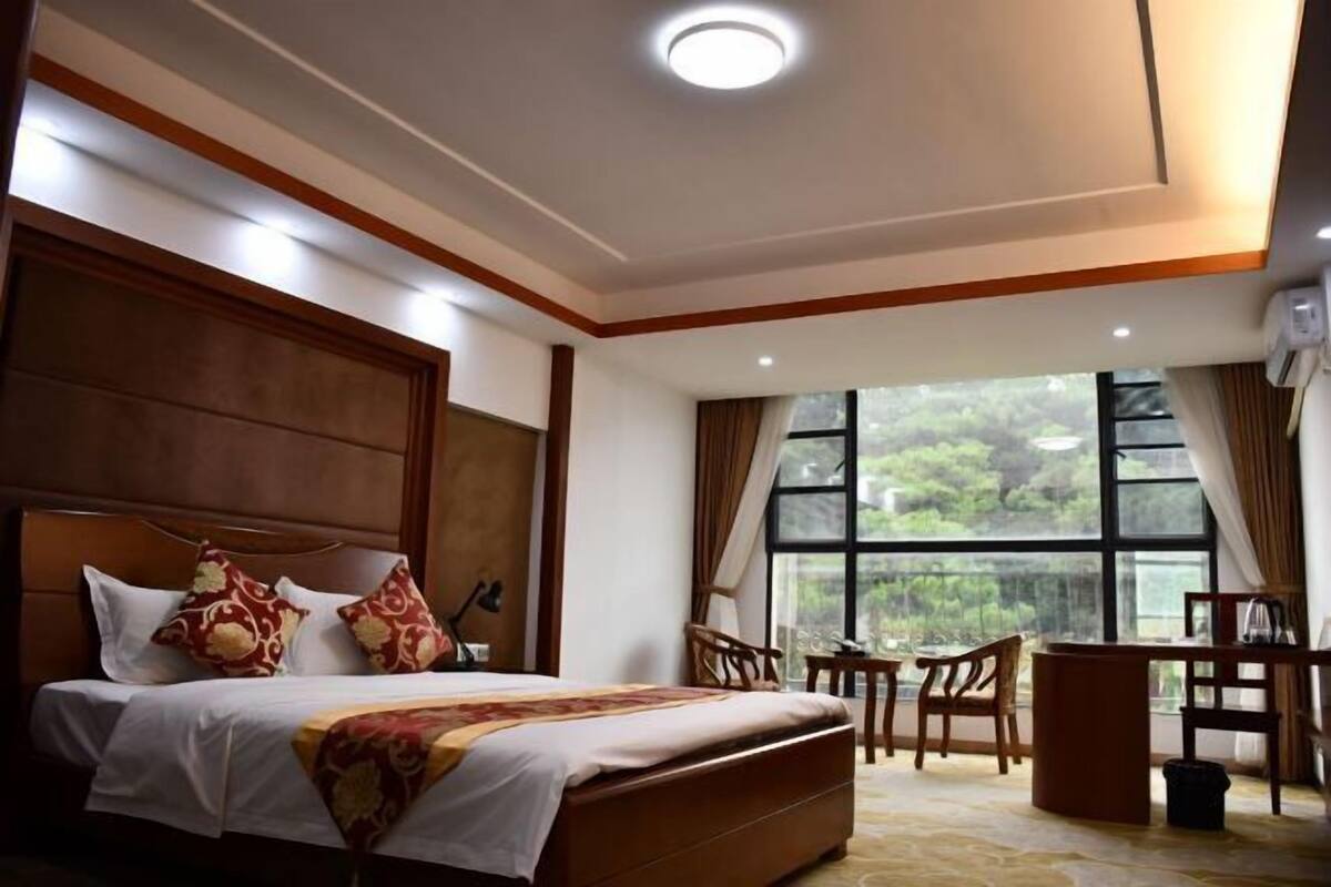 Een hotelkamer met een bed, bureau, stoelen en een groot raam met uitzicht op groen.