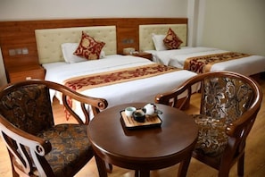Habitación de hotel con dos camas, un cabecero de madera, una mesa redonda de madera con una bandeja de té y dos sillones tapizados.