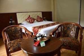 Habitación de hotel con cama, dos sillones tapizados, una mesita redonda con juego de té y un cojín decorativo.