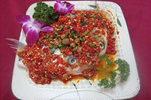 Un plato blanco con un guiso de pescado adornado con hierbas y salsa, acompañado de flores moradas y hojas verdes.