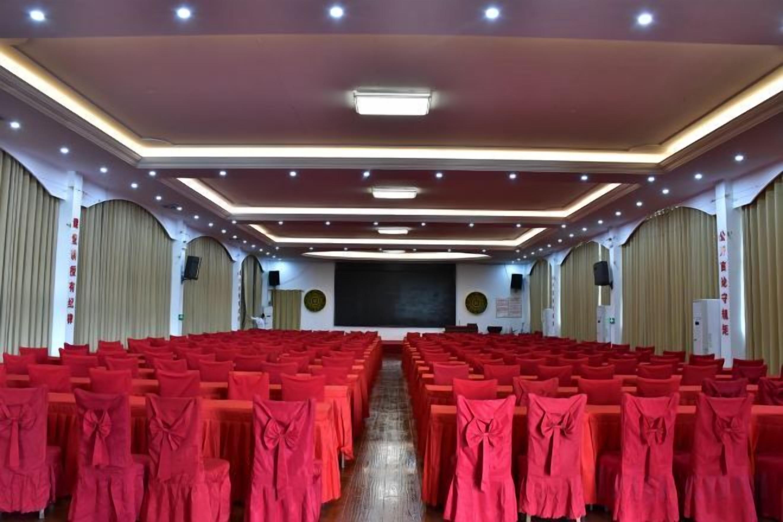 Une salle de conférence avec des rangées de tables recouvertes de nappes rouges, une scène à l’avant et un écran de projection.