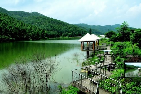 A lakeside wooden structure with a roof, surrounded by lush greenery and hills.. Nanning Natian Resort Manor