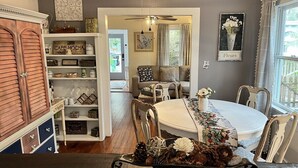 Un coin repas avec une table ronde, des chaises et un élément décoratif au centre. Un ventilateur de plafond est installé au-dessus, et une fenêtre avec des rideaux se trouve à droite.