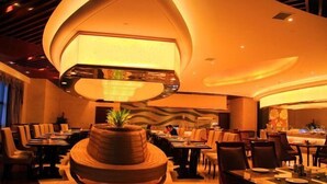 Een restaurant met een centraal zitgedeelte, met een ronde, verhoogde tafel en een grote, verlichte lamp. Tafels zijn rondom het zitgedeelte gerangschikt, en de ruimte heeft een moderne, warme sfeer.