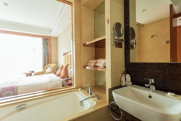 Un baño de hotel con un espejo grande, dos lavamanos, una ducha y una cama visible a través de una ventana.