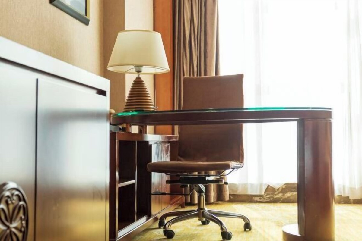 Habitación de hotel con escritorio de vidrio, silla de oficina color café, lámpara y vistas al exterior a través de una ventana con cortinas.