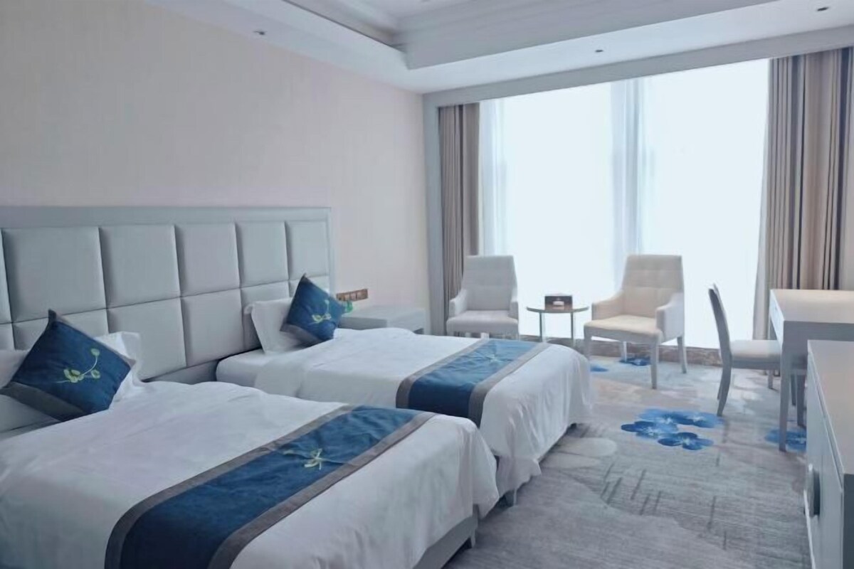 Habitación de hotel con dos camas, un ventanal grande y un área de descanso con sillas.