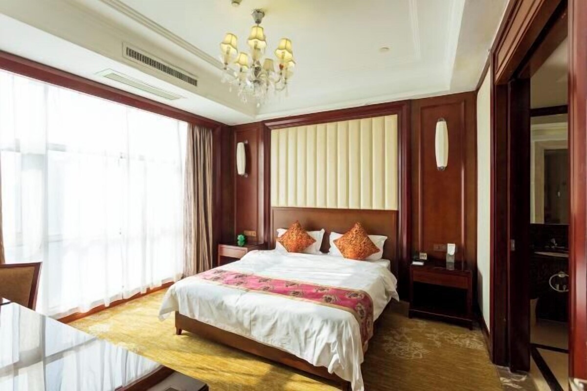 Habitación de hotel con una cama grande, un candelabro, cabecera de madera, una mesita de noche y un ventanal con cortinas.