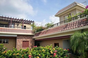 Un edificio residencial de dos pisos con un balcón, fachada de piedra y un jardín con plantas florecientes.