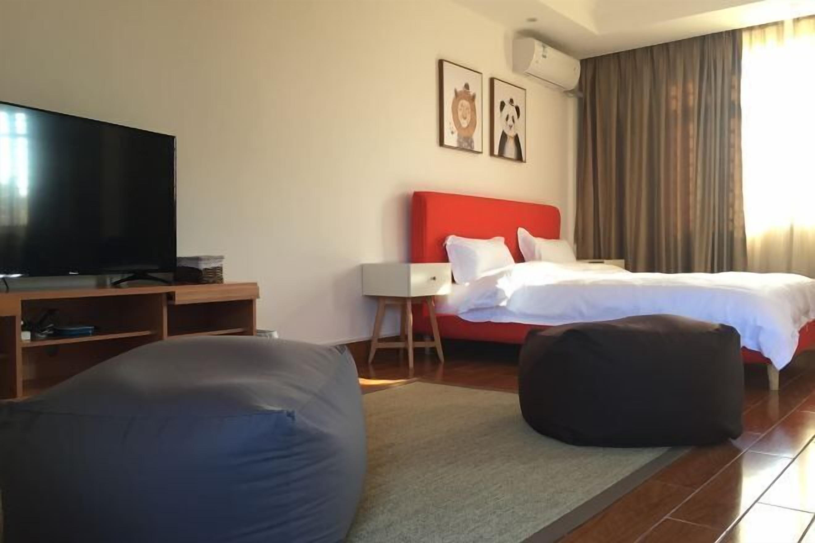 A hotel room with a flat-screen TV, a red headboard bed, two framed pictures on the wall, a large bean bag chair, and a wooden desk.