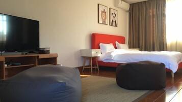 A hotel room with a flat-screen TV, a red headboard bed, two framed pictures on the wall, a large bean bag chair, and a wooden desk.