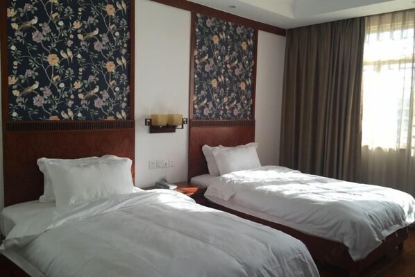 Habitación de hotel con dos camas, un gran tapiz floral en la pared, una mesita de noche con lámpara y una ventana con cortinas.