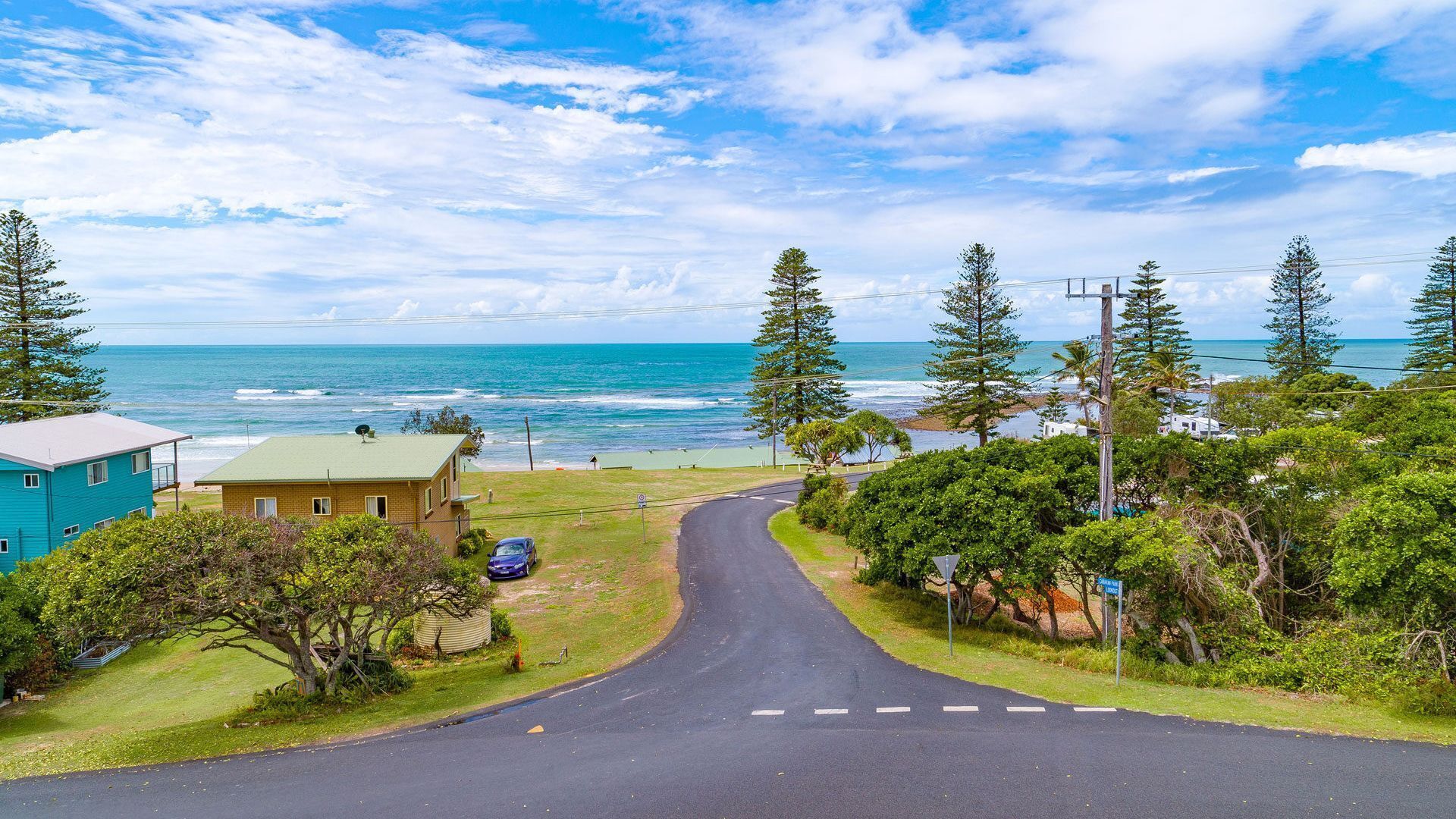A coastal road leading to a beach with houses on one side and a clear view of the ocean.