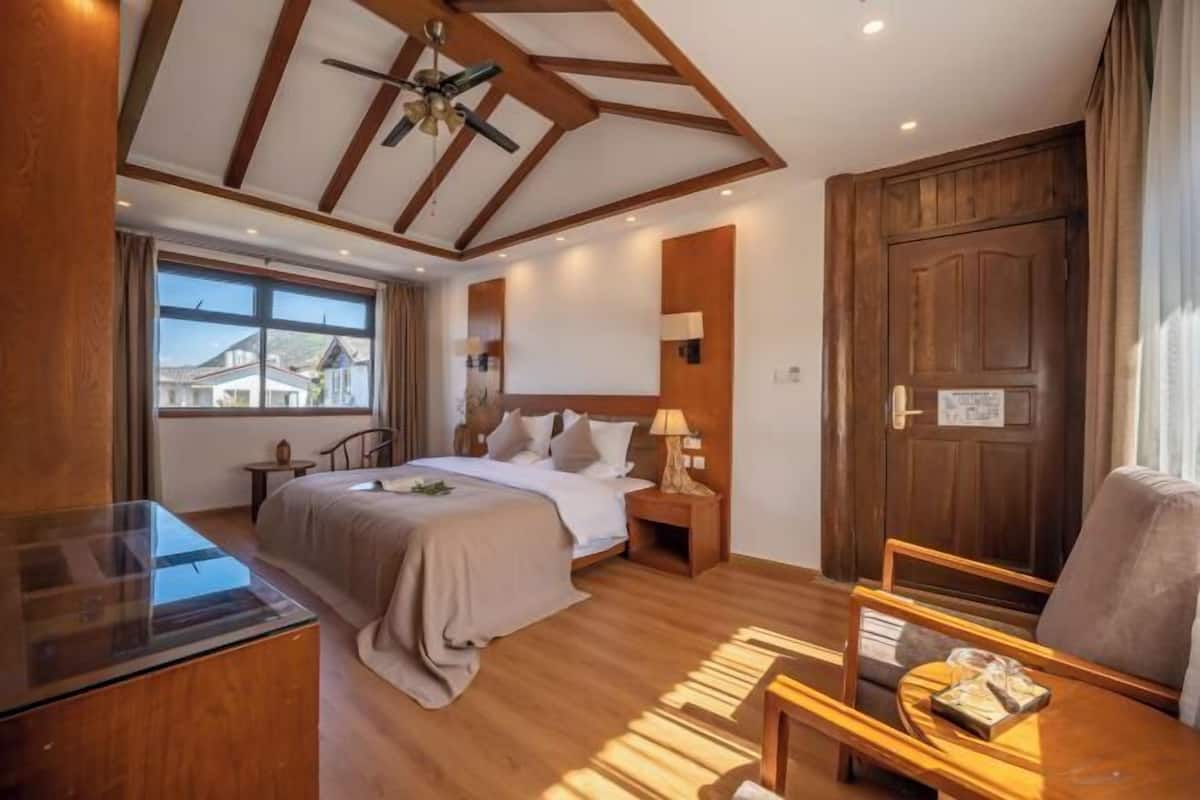 A hotel room with a bed, bedside table, lamp, wooden floor, ceiling fan, and a view of buildings outside.