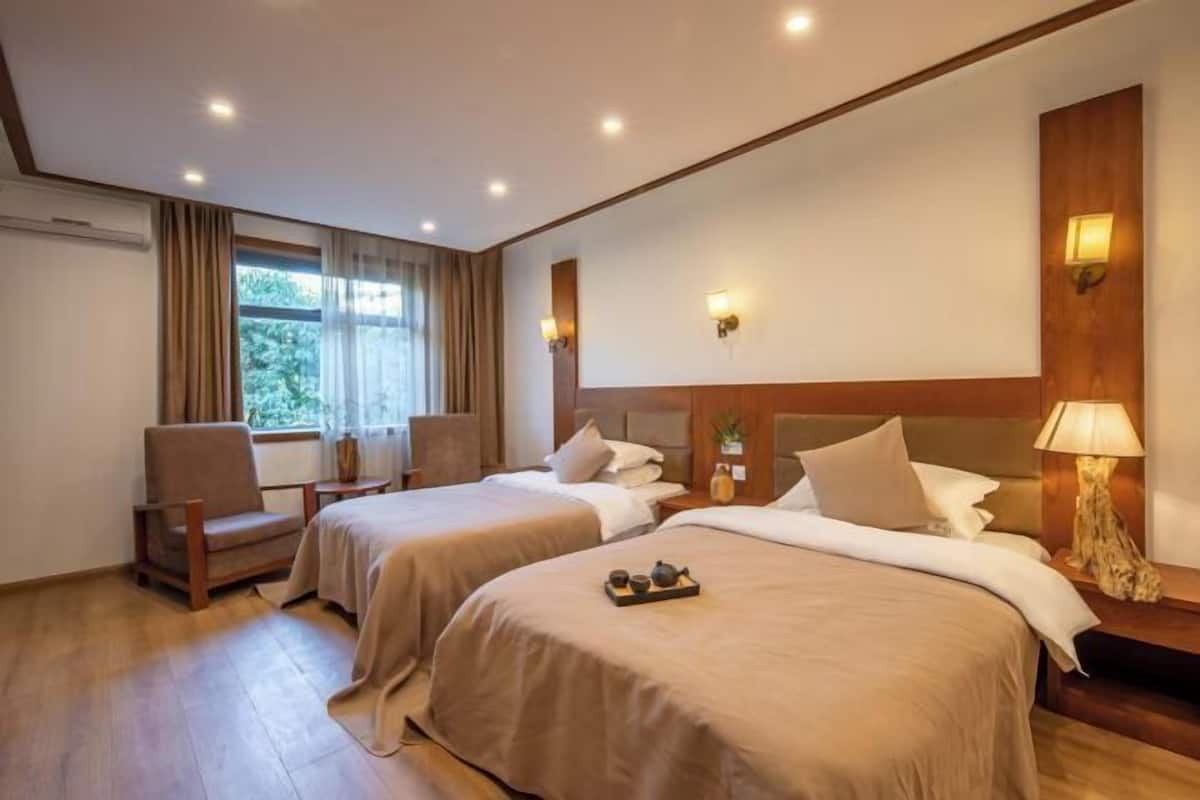 A hotel room with two beds, a chair, a window with curtains, and wall-mounted lamps.