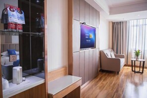 Habitación de hotel moderna con televisión de pantalla plana montada en la pared, una zona de estar con sofá y sillones, una mesita y un separador de vidrio con una cafetera y un estante con agua embotellada.