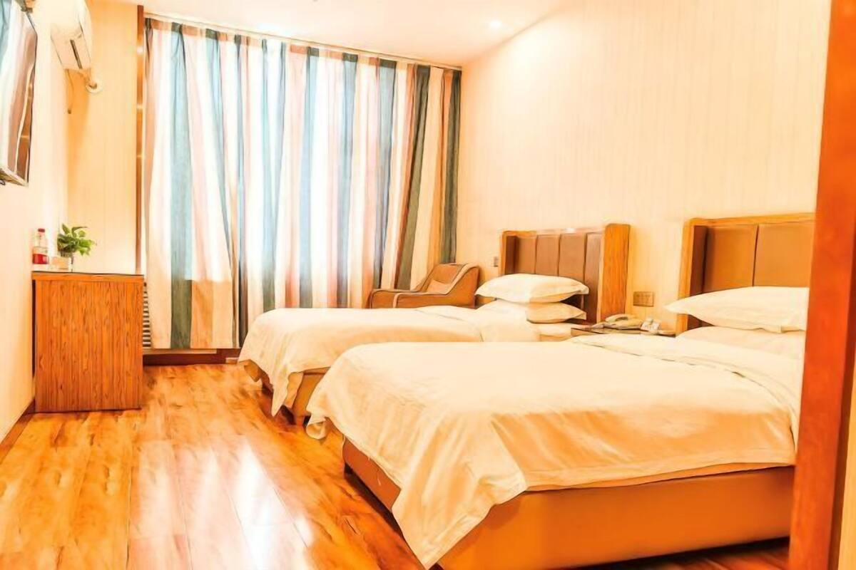 A hotel room with two beds, a wooden floor, a desk with a chair, a window with curtains, and a wall-mounted phone.