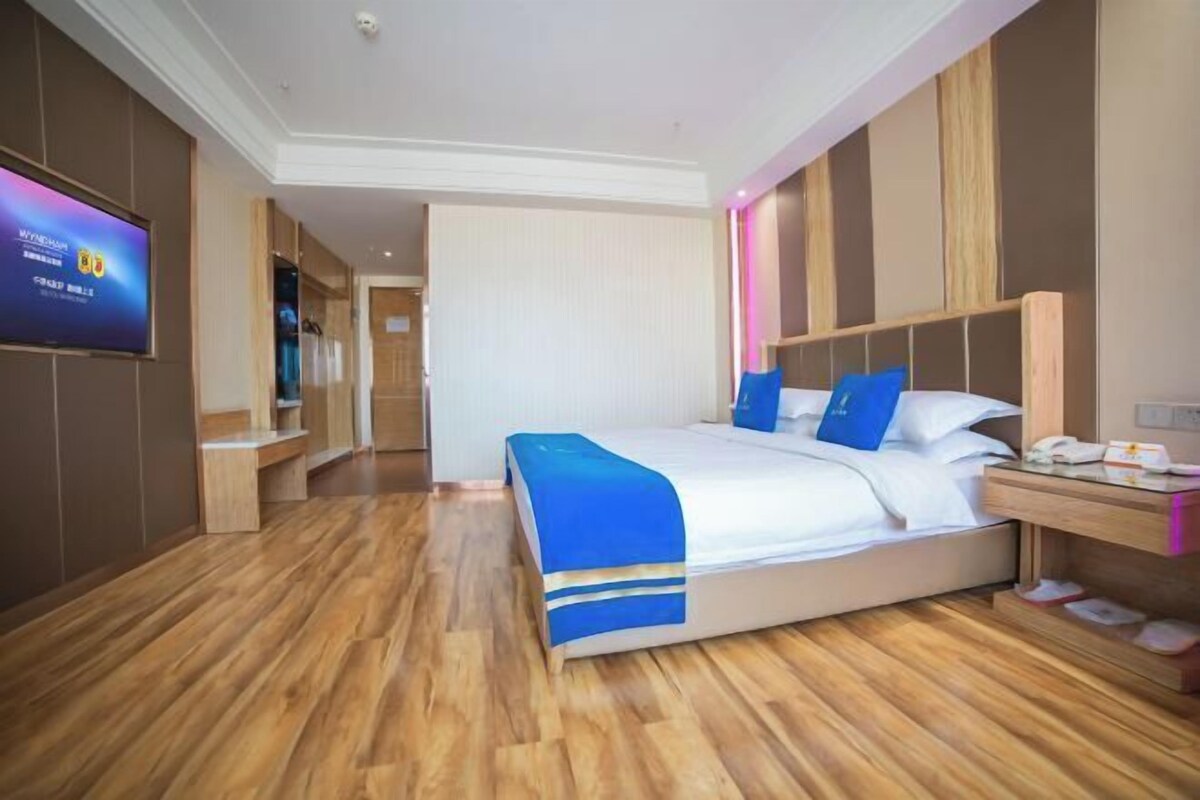 Habitación de hotel con una cama grande, piso de madera, un televisor en la pared y una mesita de noche con un teléfono y pantuflas.