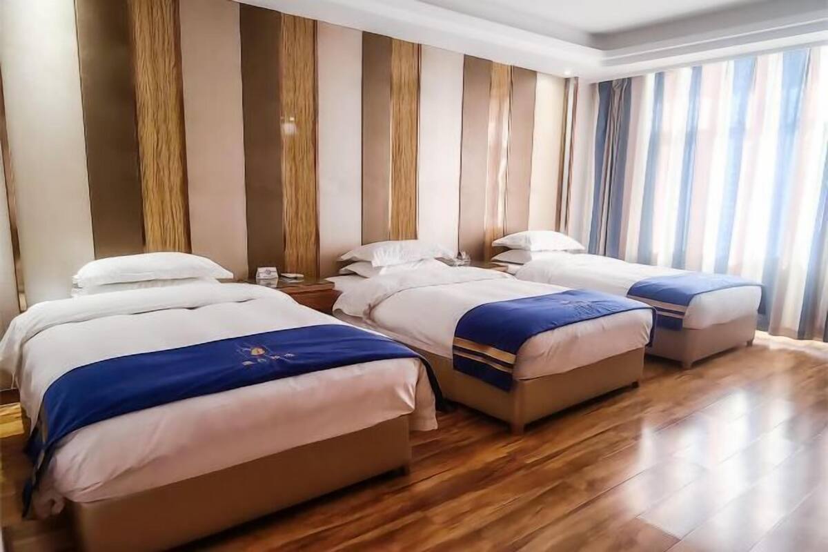 A hotel room with four beds arranged in a row, wooden flooring, and a large window with curtains.