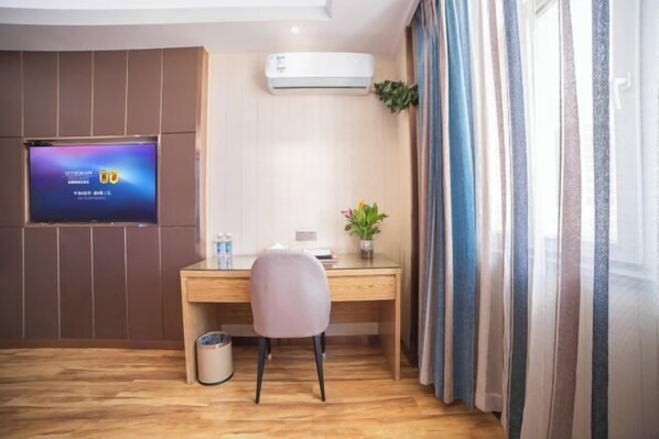 A hotel room with a wooden desk, a chair, a television mounted on the wall, a trash bin, and a window with curtains.