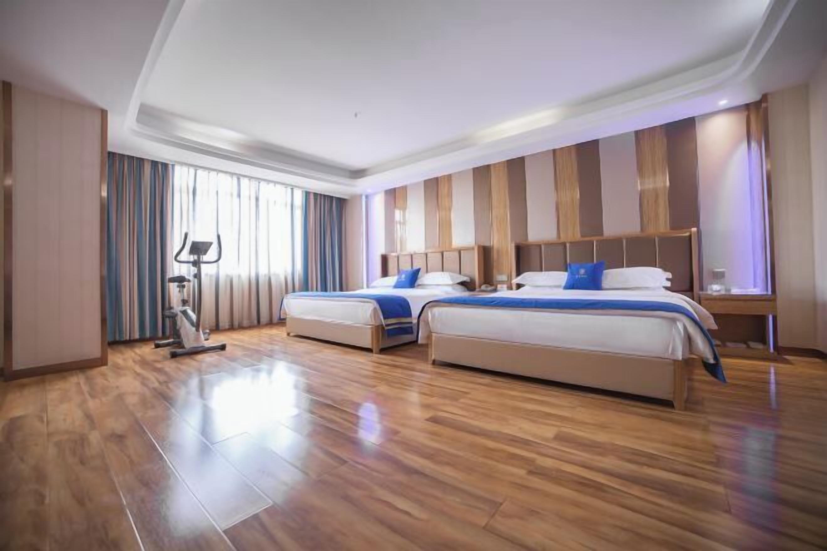 Habitación de hotel con dos camas, una cinta caminadora y un ventanal con cortinas.