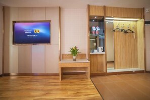 Zona de recepción de un hotel con un televisor mostrando el logotipo de Wyndham, un banco de madera con una planta en maceta y un armario de madera con espejo y ganchos.