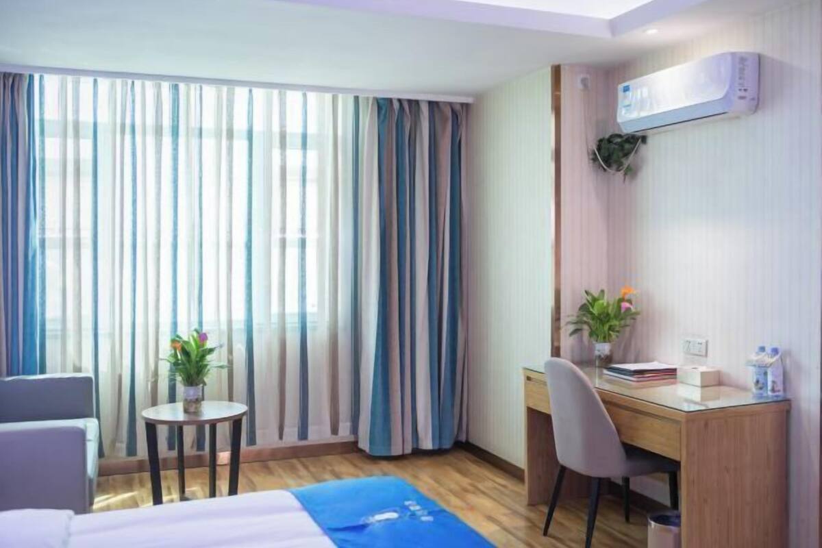 A hotel room with a bed, a desk, a chair, a small table with a plant, and a window with curtains.