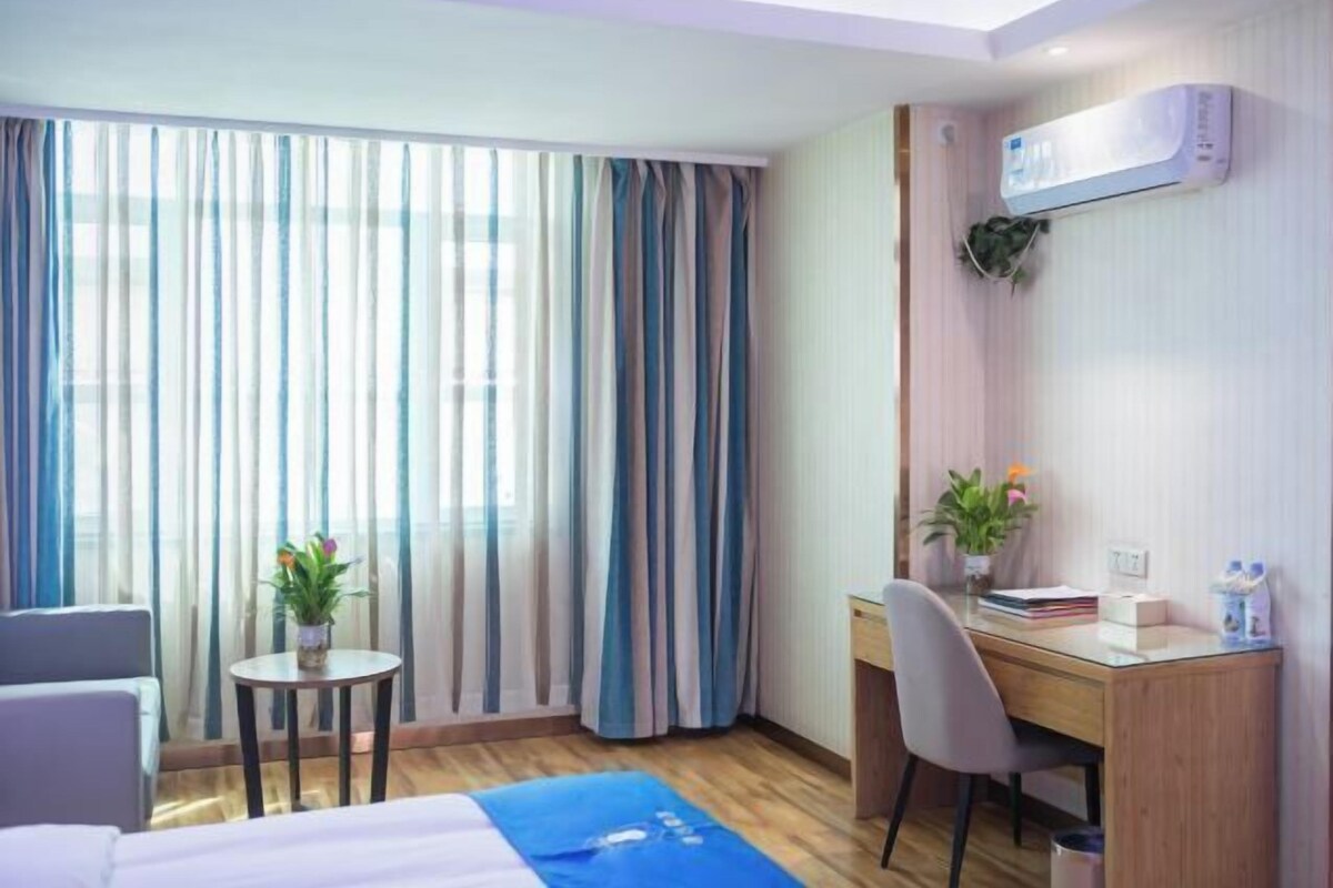 Habitación de hotel con cama, escritorio, silla, una mesita con una planta y ventana con cortinas.